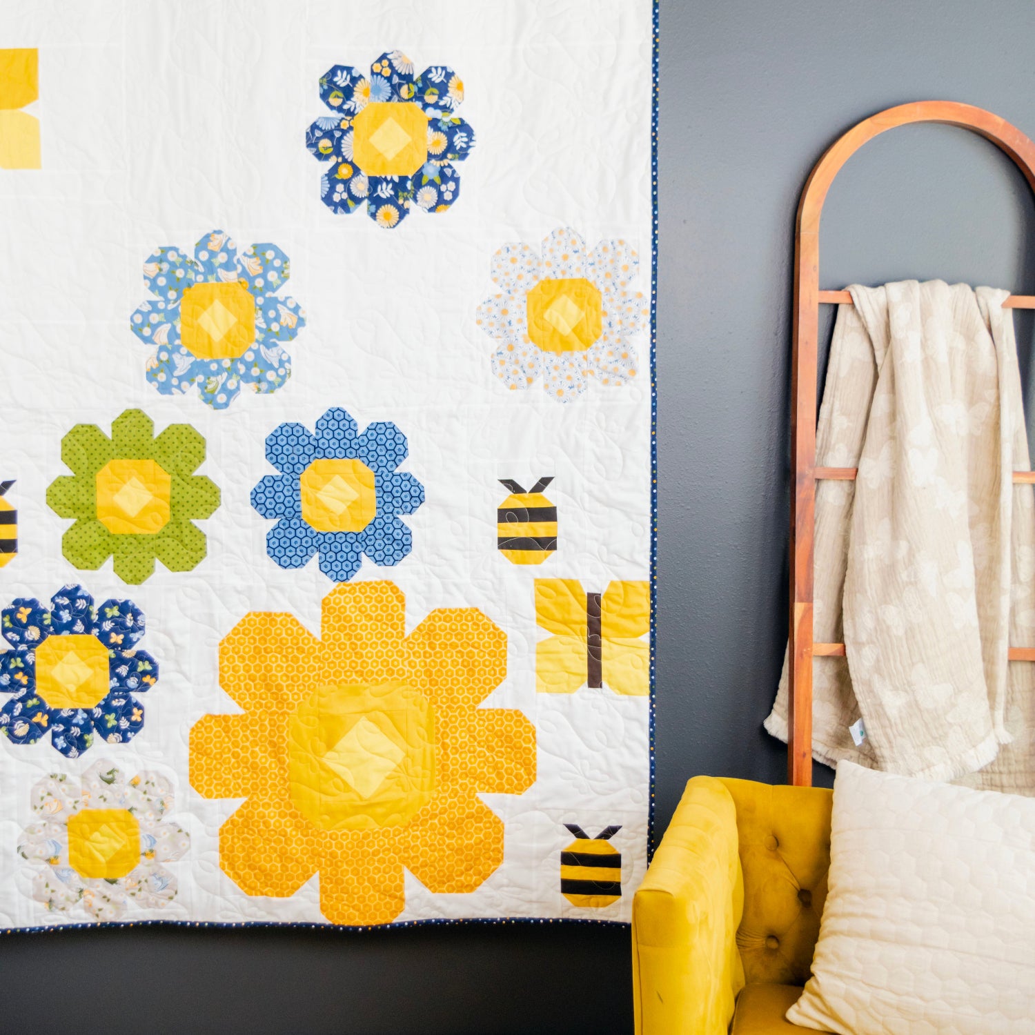 A white quilted wall hanging or blanket with large yellow and blue flower motifs, green flower accents, and small bumblebee designs, displayed in a styled room next to a yellow chair and a wooden floor.