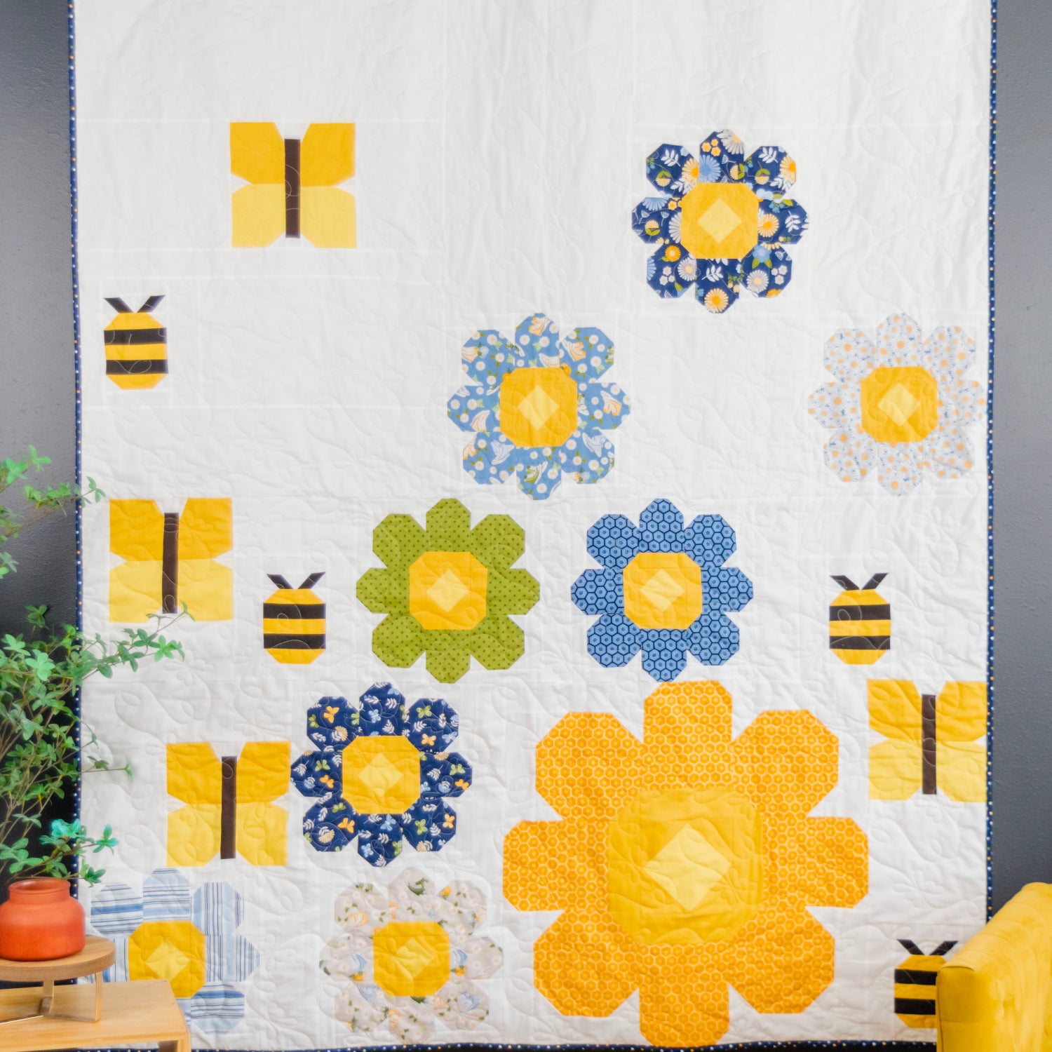 A quilted wall hanging or blanket featuring a whimsical bee and flower pattern in yellow, blue, and green on a white background, styled in a bedroom setting next to a yellow chair and a plant.