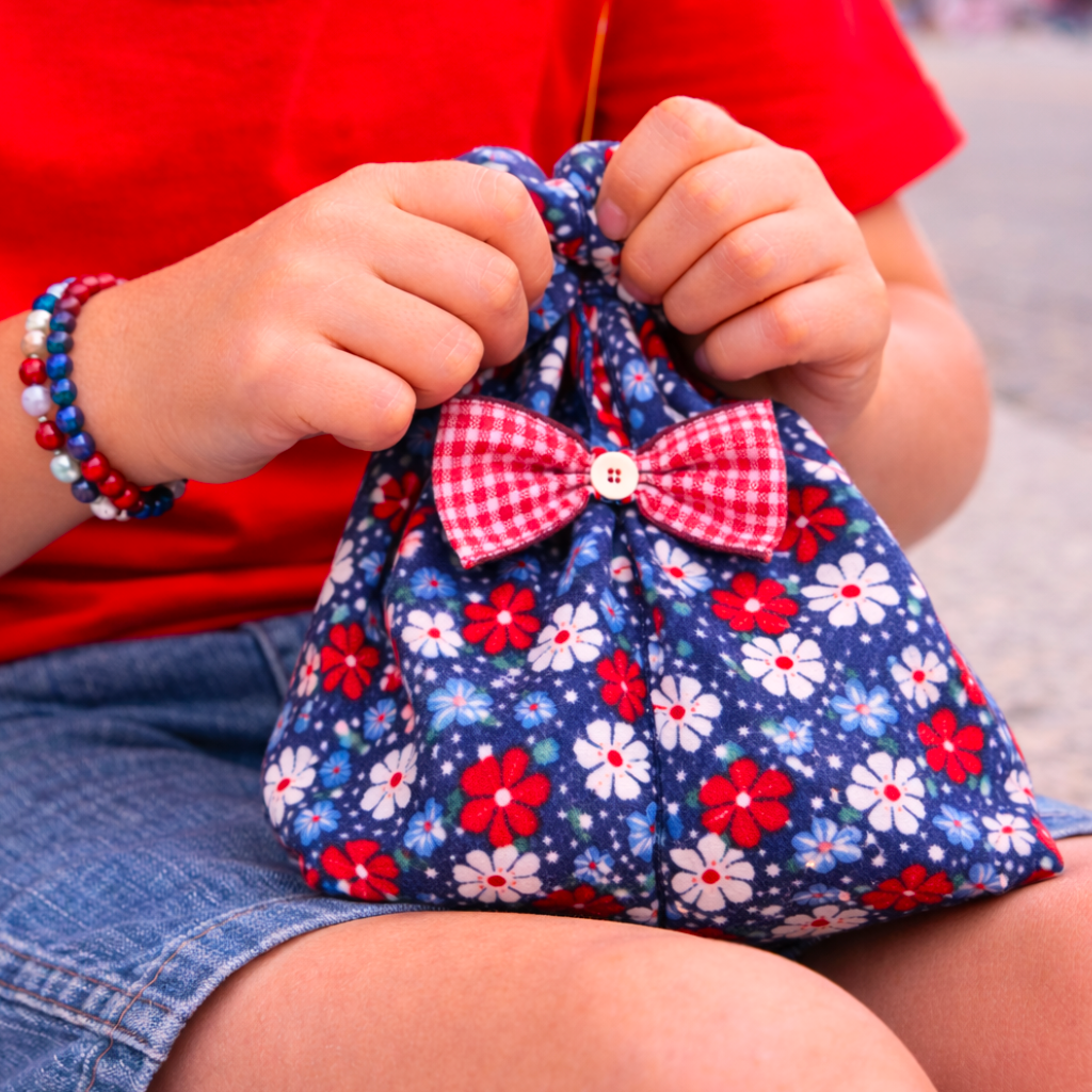 Child holding a small drawstring bag with a red, white, and blue floral pattern and bow.