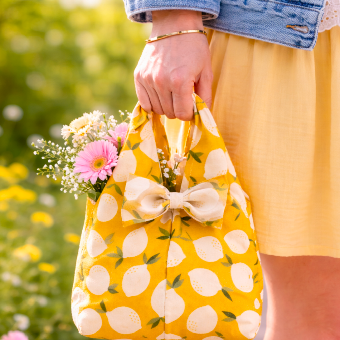 Person holding a yellow reusable bag with lemon pattern filled with flowers, outdoors.