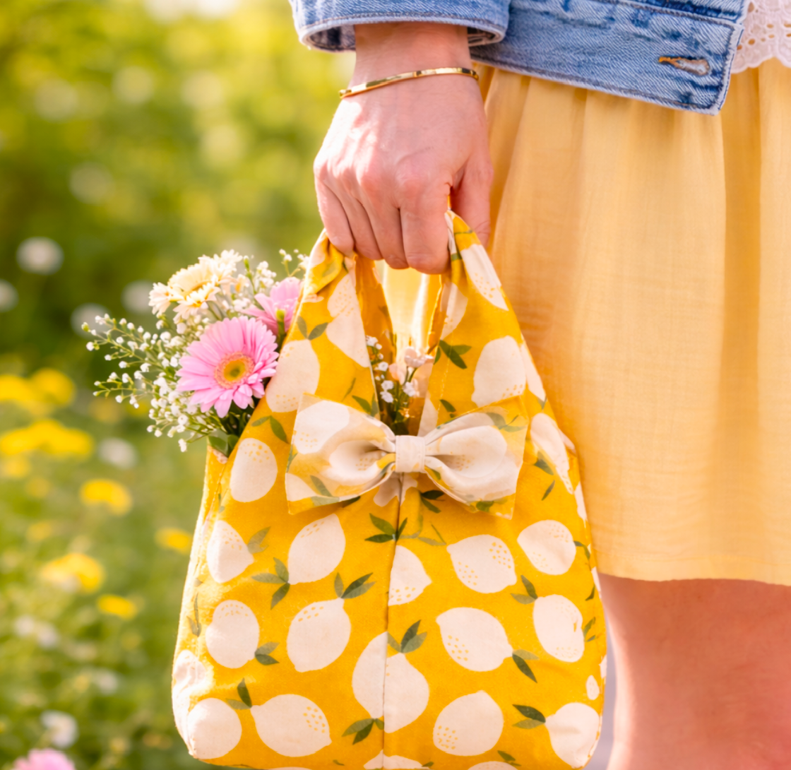 Person holding a yellow reusable bag with lemon pattern filled with flowers, outdoors.