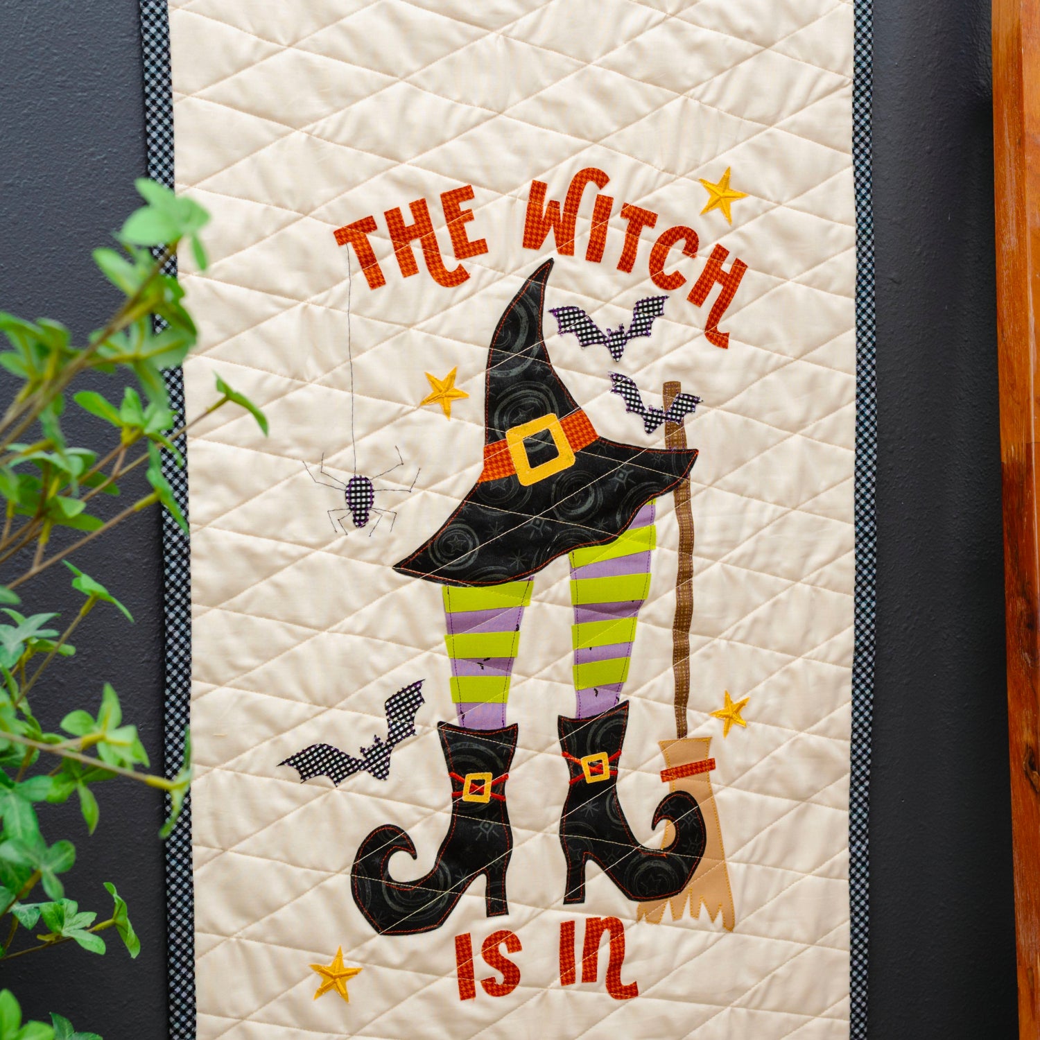 A vertical quilted wall hanging or tapestry with a Halloween witch design, shown hanging on a dark wall next to a wooden ladder holding another quilt. The main design features the words 'THE WITCH IS IN' in orange, a black witch hat, green-and-yellow striped legs, black boots, bats, and stars.