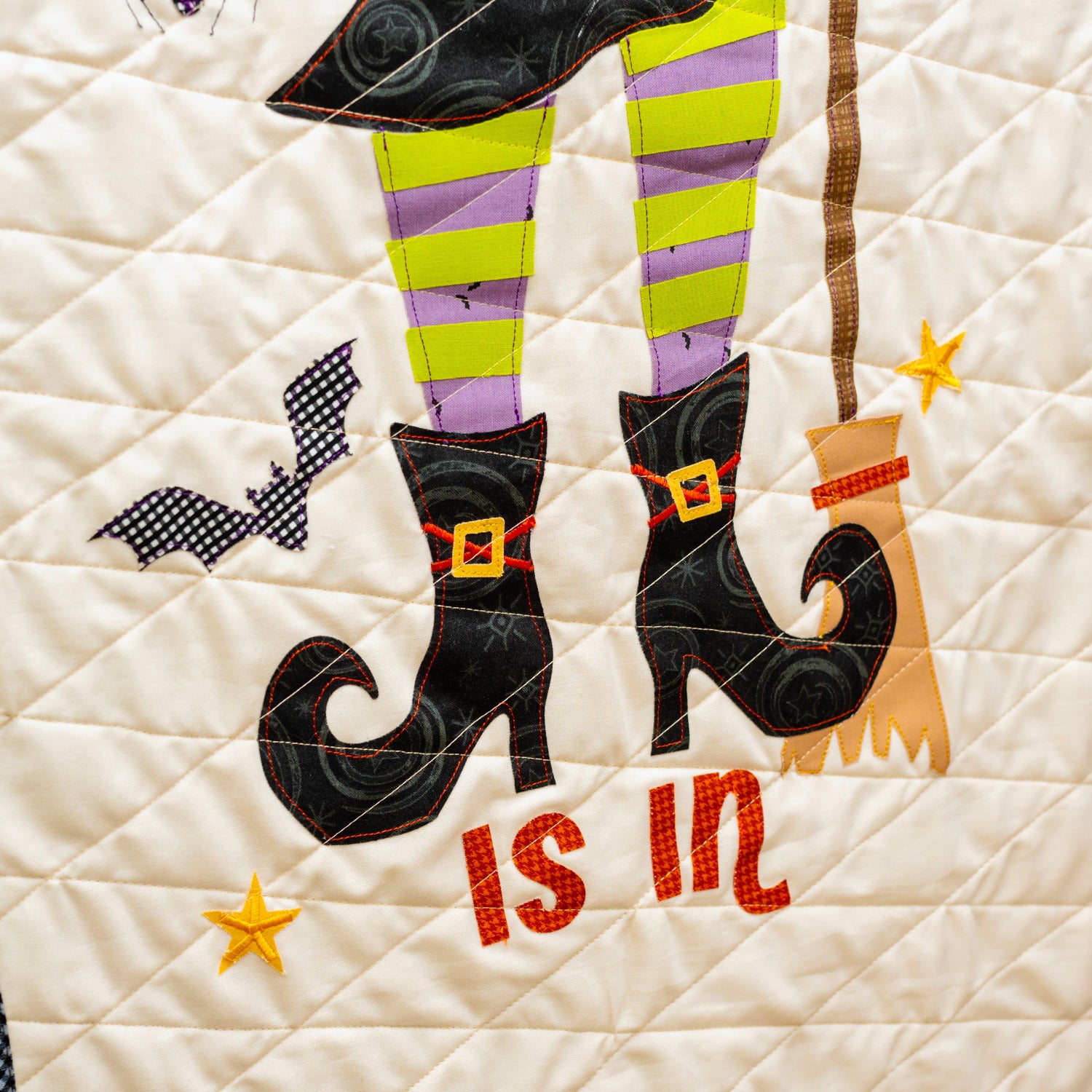 A close-up of a quilted fabric wall hanging or panel with a Halloween design. The cream-colored quilt features applique legs in black and purple striped stockings, black witch boots with gold buckles, a broom, stars, and the words 'is in' in red.