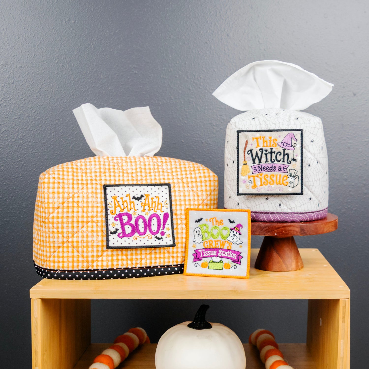 Two Halloween-themed tissue box covers displayed on a wooden shelf against a dark gray wall. One cover is orange and white gingham with a black polka-dot trim and a 'Don't Hype BOO!' patch, and the other is white speckled with a black polka-dot border and a 'This Witch Must Be Obsessed' patch.