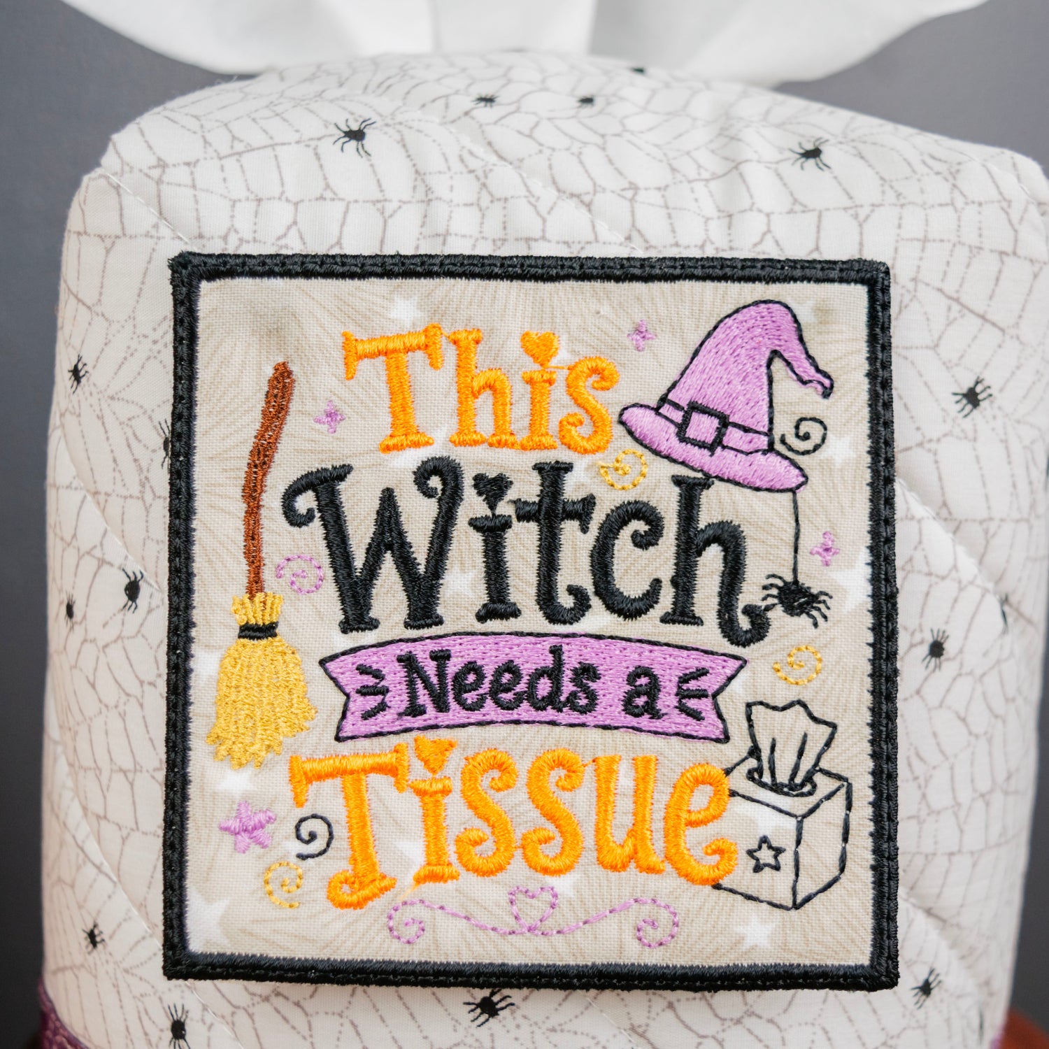 A close-up of a Halloween-themed fabric tissue box cover with embroidered text reading 'This Witch Needs a Tissue.' The cover is off-white with a black spiderweb pattern and features a central embroidered patch with a purple witch hat, broom, spiders, and orange and black lettering.