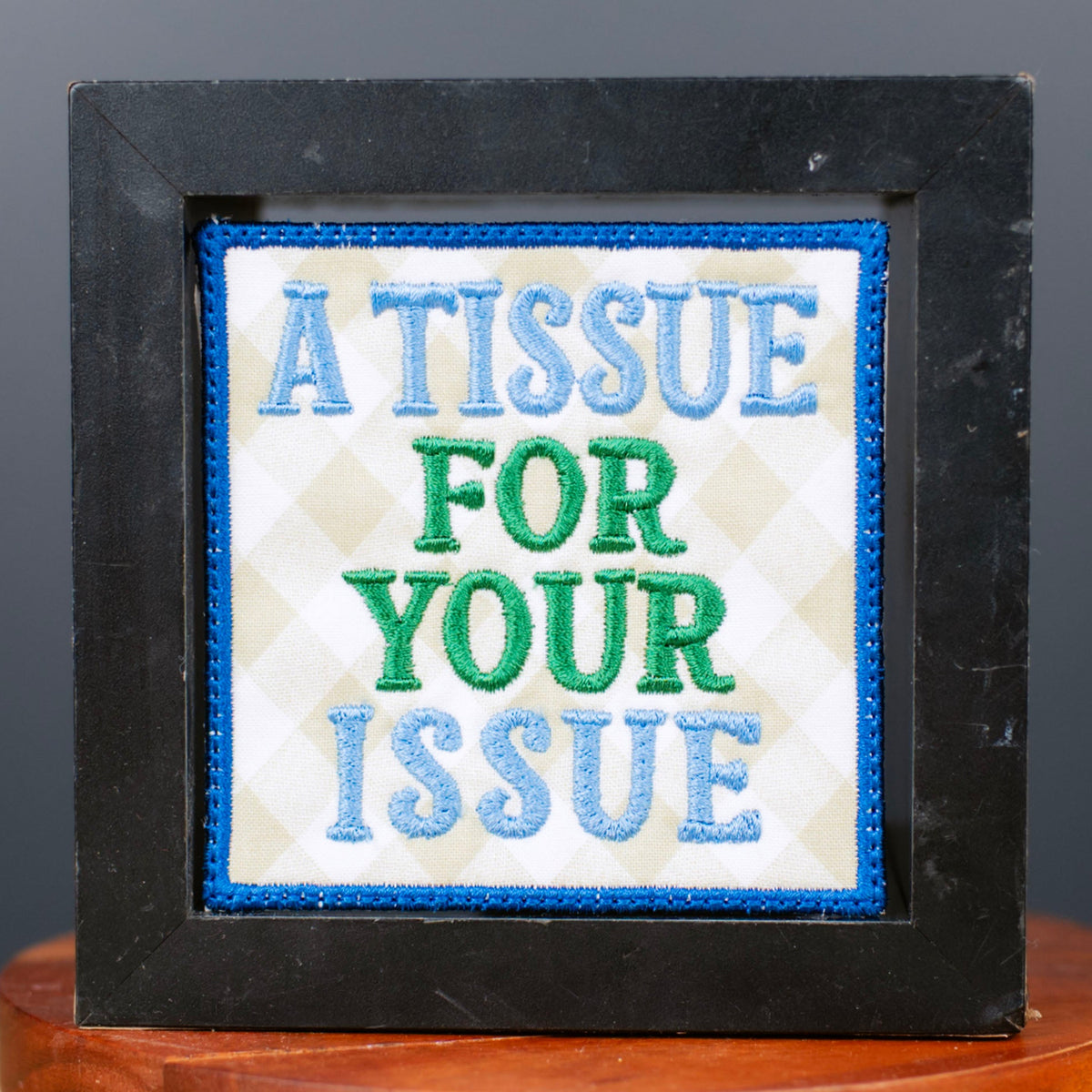 A square black-framed novelty sign with a white center panel reading 'A TISSUE FOR YOUR ISSUE' in blue and green text, displayed on a small wooden stand against a dark gray background.