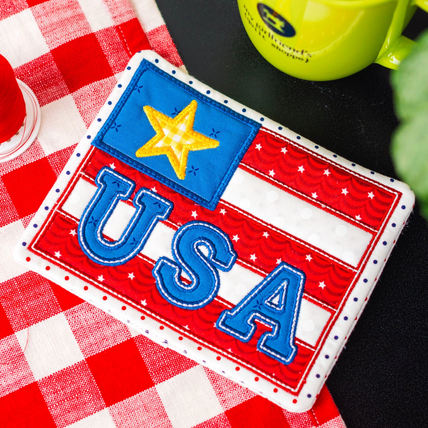 USA-themed patch on a red and white checkered tablecloth with a green mug and red flower in the background.