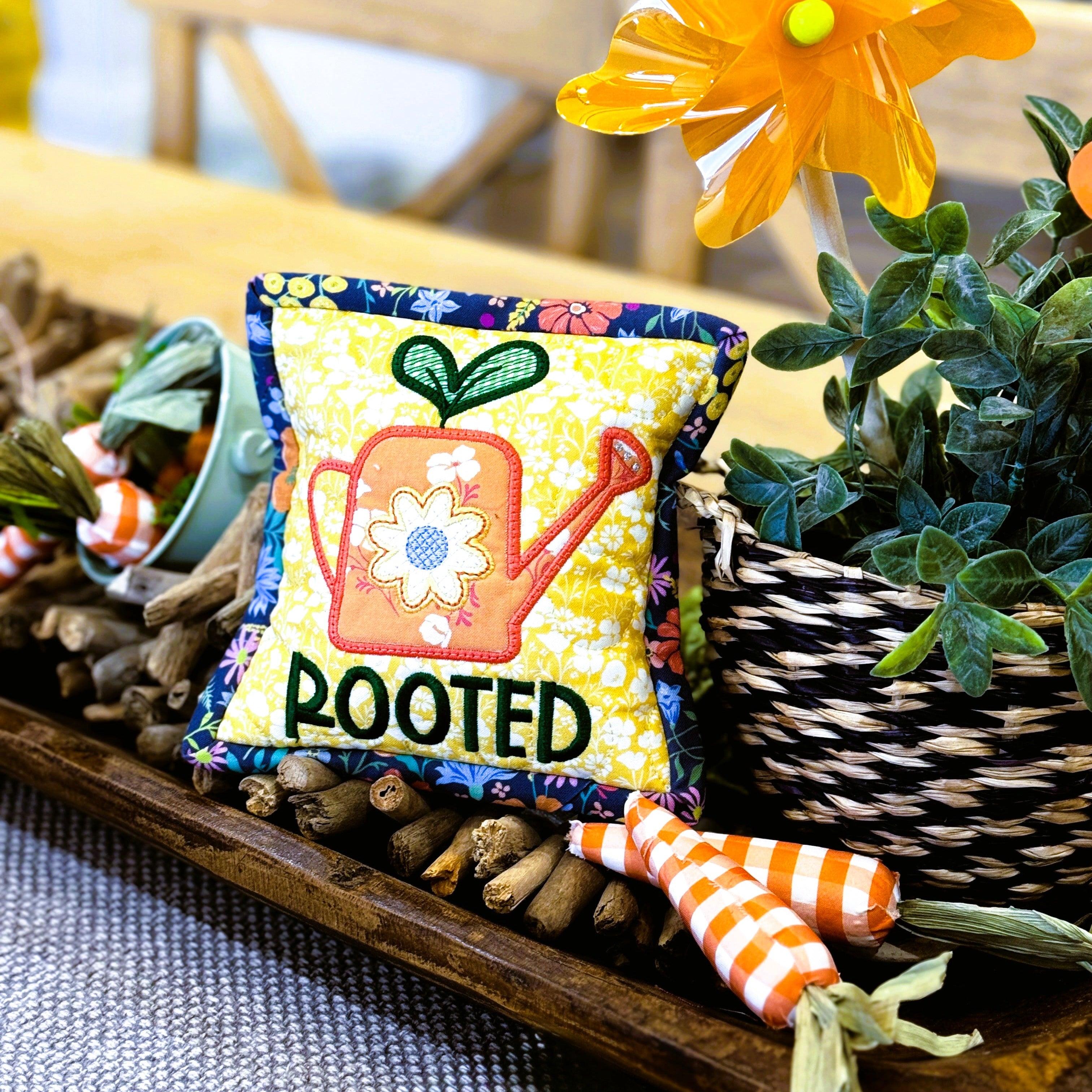 Decorative pillow with 'ROOTED' text and a sunflower on a table with plants and carrots.