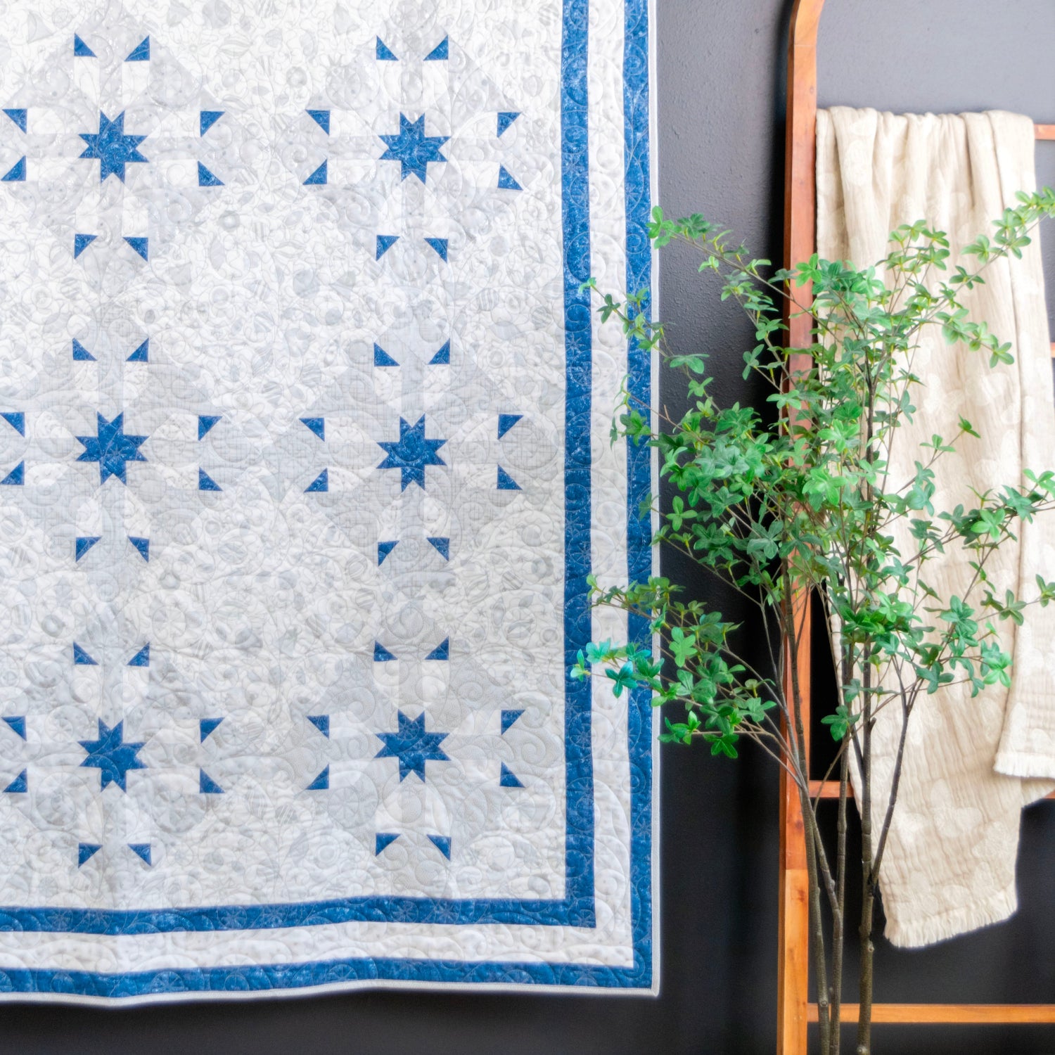 A blue and white quilted wall hanging with a geometric star pattern, displayed next to a wooden mirror and a potted green plant against a dark gray wall.