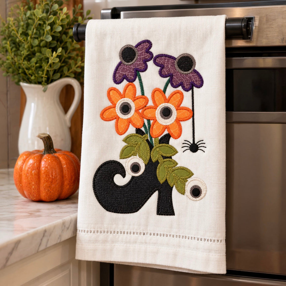 A cream-colored kitchen towel with an embroidered autumn design of orange and purple flowers, green leaves, a black ghost, and a spider, hanging on a black oven handle in a kitchen setting.