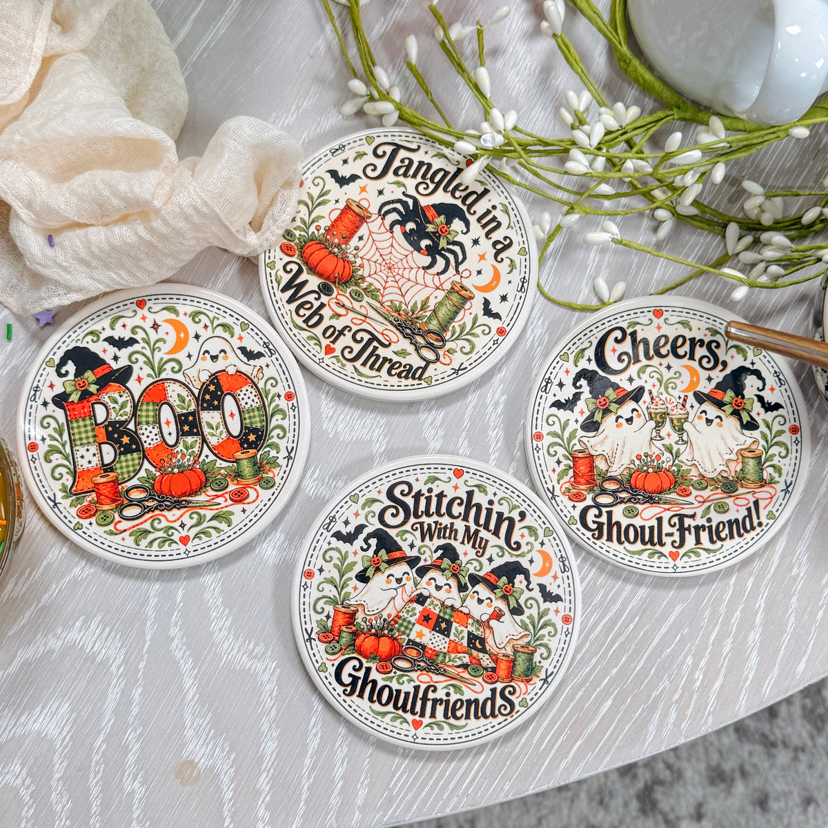 Four round coasters with detailed Halloween illustrations and text, arranged on a white textured tabletop with decorative props including flowers, a mug, and soda cans.
