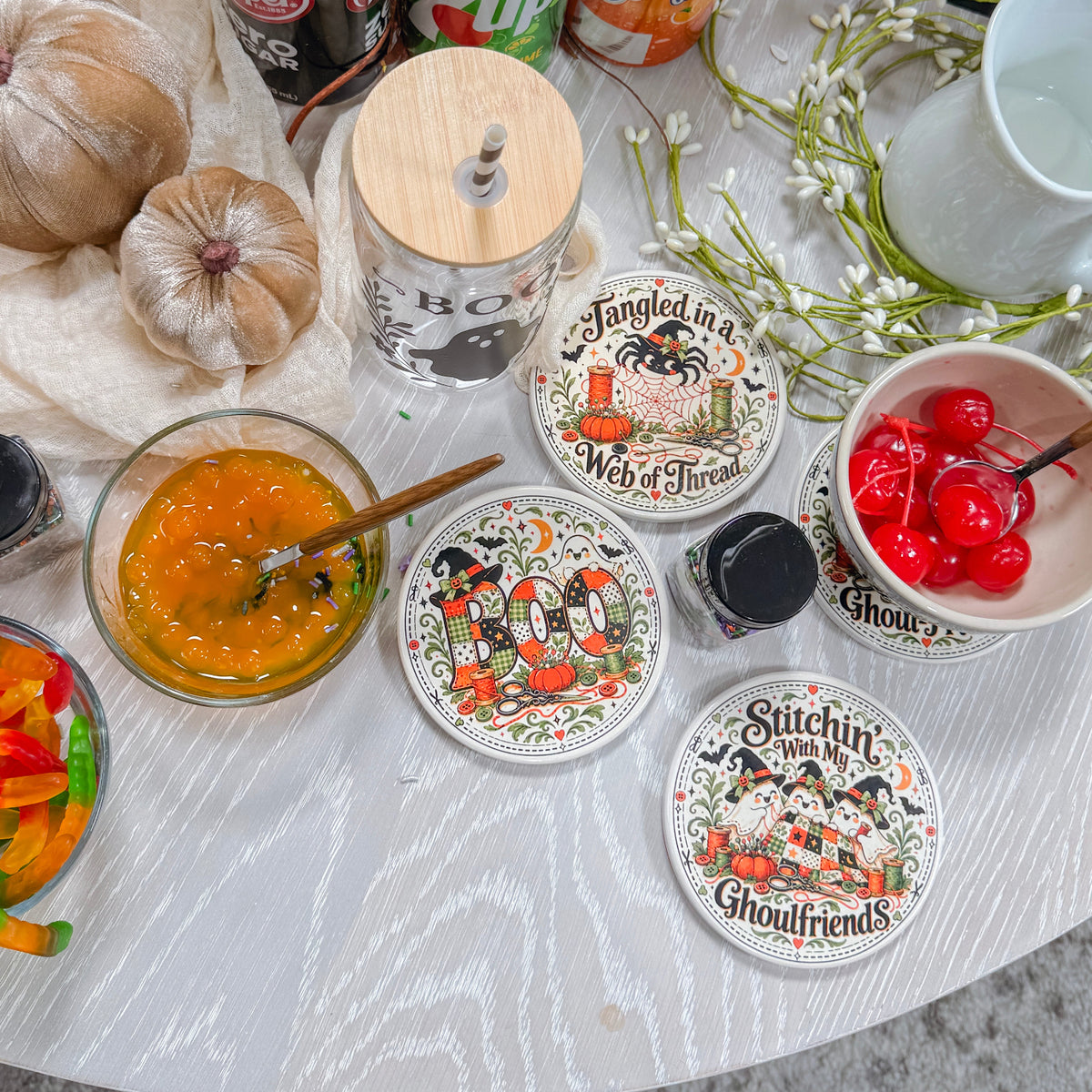 A styled tabletop scene with multiple beverage cans, a glass bottle with a wooden lid, and several round coasters featuring whimsical illustrated designs. There are also bowls of food, garlic bulbs, flowers, and a white pitcher on a light wood surface.