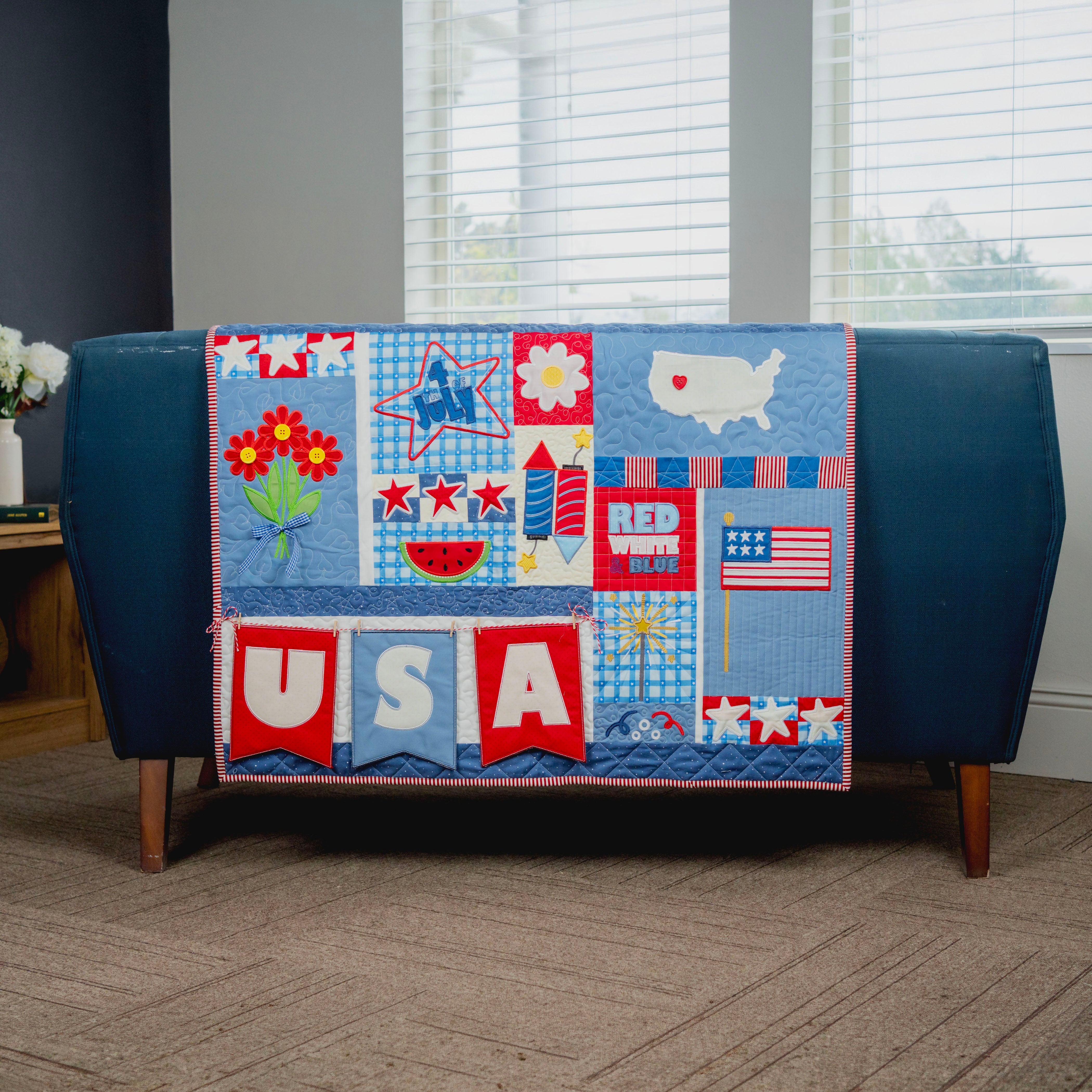 Colorful quilt with patriotic designs draped over a blue couch in a room with windows.