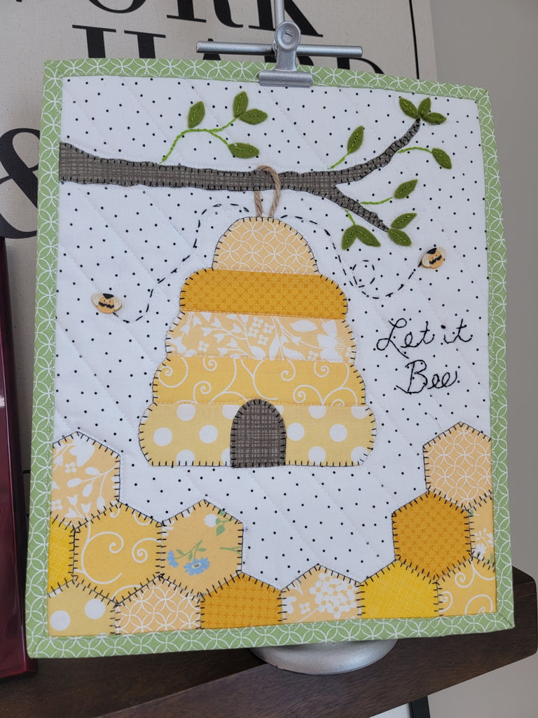 A miniature quilt featuring a beehive and the words "Let it Bee".