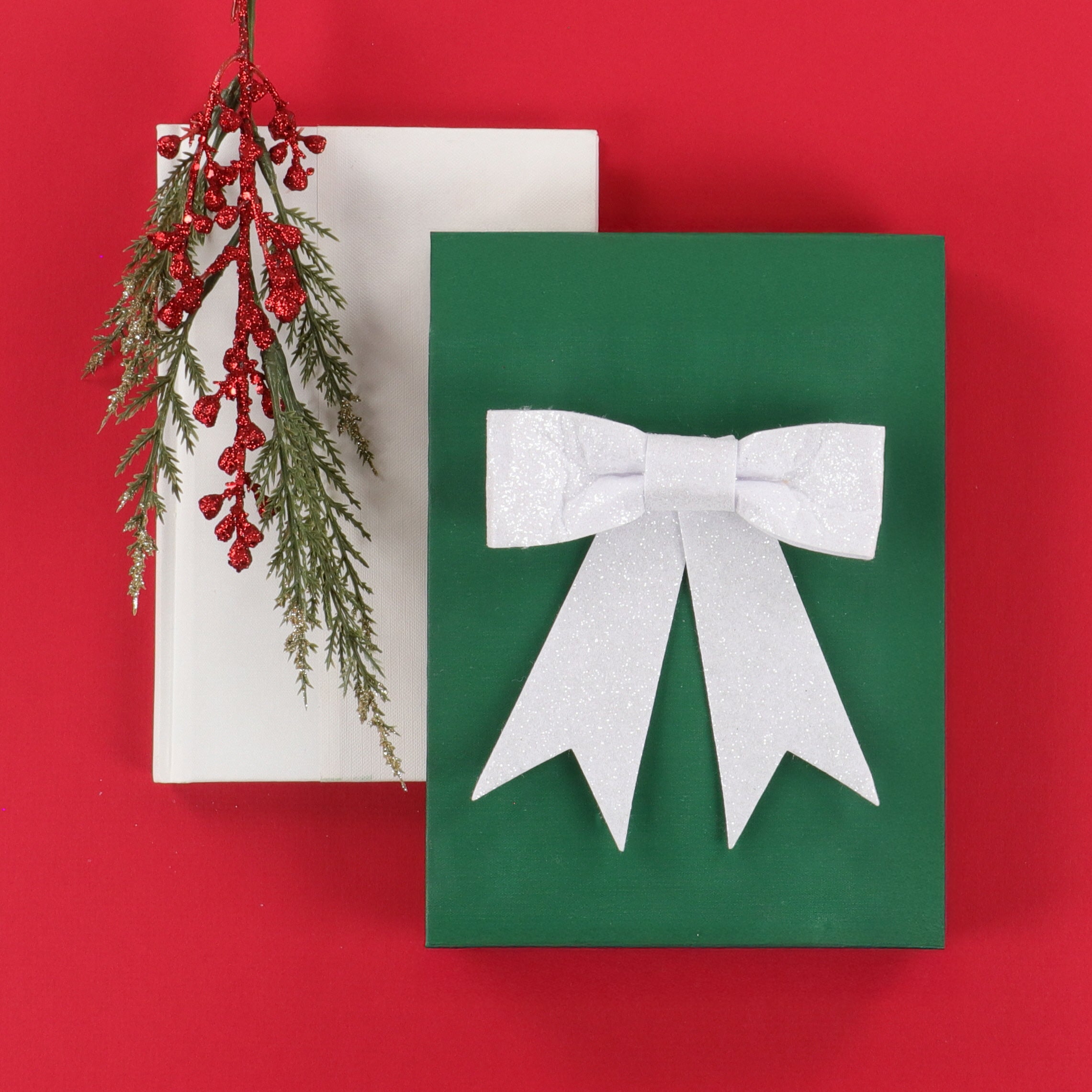 A green rectangular gift box with a large white glitter bow on the front, placed on a red background with a white box and a hanging red berry garland behind it.