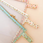 Three clear zippered pouches with floral fabric trim in pastel pink, mint green, and white, arranged diagonally on a solid pale peach background.