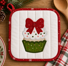 A quilted white pot holder with a red border and an applique of a cupcake topped with a red bow, shown on a wooden table with holiday decor.