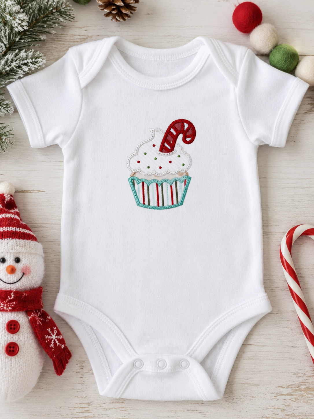 A white short-sleeve baby bodysuit laid flat on a light wood surface, featuring a cupcake graphic with white frosting, red and green sprinkles, and a red-and-white candy cane. The bodysuit is styled with holiday props including a snowman plush, candy canes, pinecones, and pom-poms.
