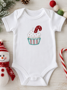 A white short-sleeve baby bodysuit laid flat on a light wood surface, featuring a cupcake graphic with white frosting, red and green sprinkles, and a red-and-white candy cane. The bodysuit is styled with holiday props including a snowman plush, candy canes, pinecones, and pom-poms.