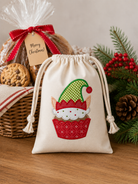 A cream fabric drawstring gift bag with an embroidered cupcake elf design, styled on a wooden table with holiday greenery, a wicker basket, and a wrapped cookie gift bag in the background.