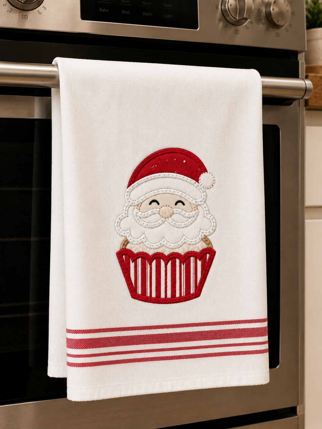 A white kitchen towel hanging from a stainless steel oven handle, featuring an embroidered Santa Claus face inside a red cupcake with red stripes at the bottom.