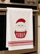 A white kitchen towel hanging from a stainless steel oven handle, featuring an embroidered Santa Claus face inside a red cupcake with red stripes at the bottom.