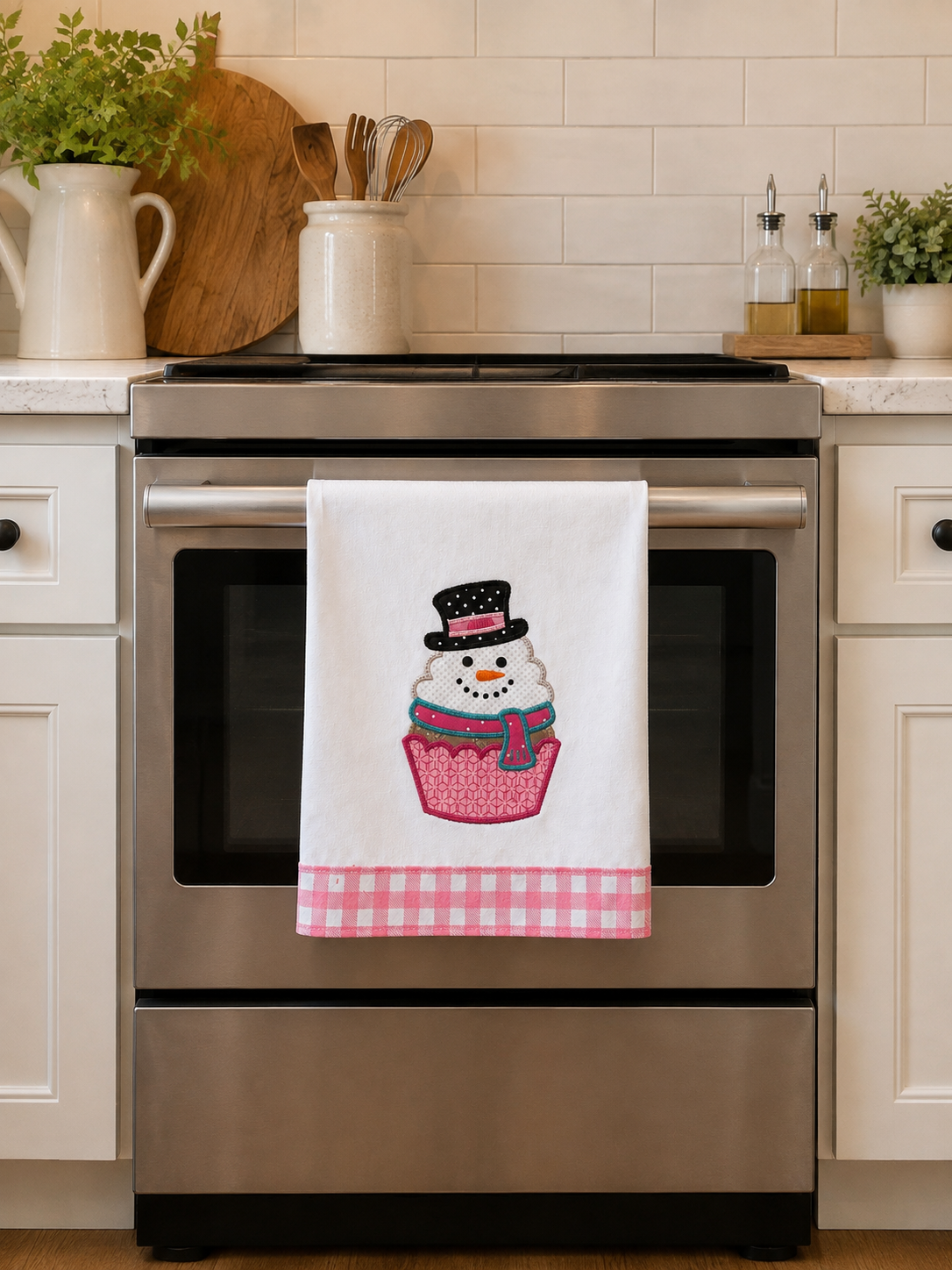 A white kitchen towel hanging on a stainless steel oven handle, featuring a cupcake with a snowman wearing a black top hat and a pink gingham bottom border. The scene is set in a bright, modern kitchen with white cabinets and a tiled backsplash.
