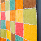 A colorful patchwork rag quilt hanging on a dark gray wall next to a wooden ladder, featuring square fabric blocks in orange, yellow, green, red, and brown.