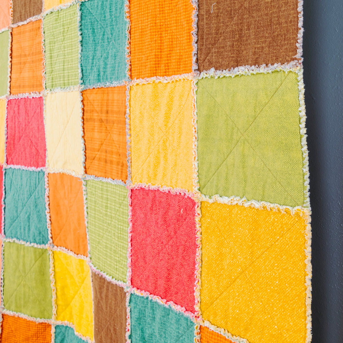 A colorful patchwork rag quilt hanging on a dark gray wall next to a wooden ladder, featuring square fabric blocks in orange, yellow, green, red, and brown.