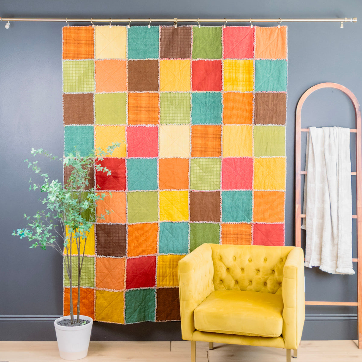 A colorful patchwork quilt hanging on a metal rod in a styled room, with a yellow armchair, a potted plant, and a wooden hanger with a white towel against a dark gray wall.