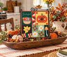 A framed autumn-themed quilted wall hanging with the word 'Grateful' and colorful patchwork motifs including acorns, a leaf, daisies, mushrooms, and pumpkins, displayed on a dining table in a decorated fall setting.