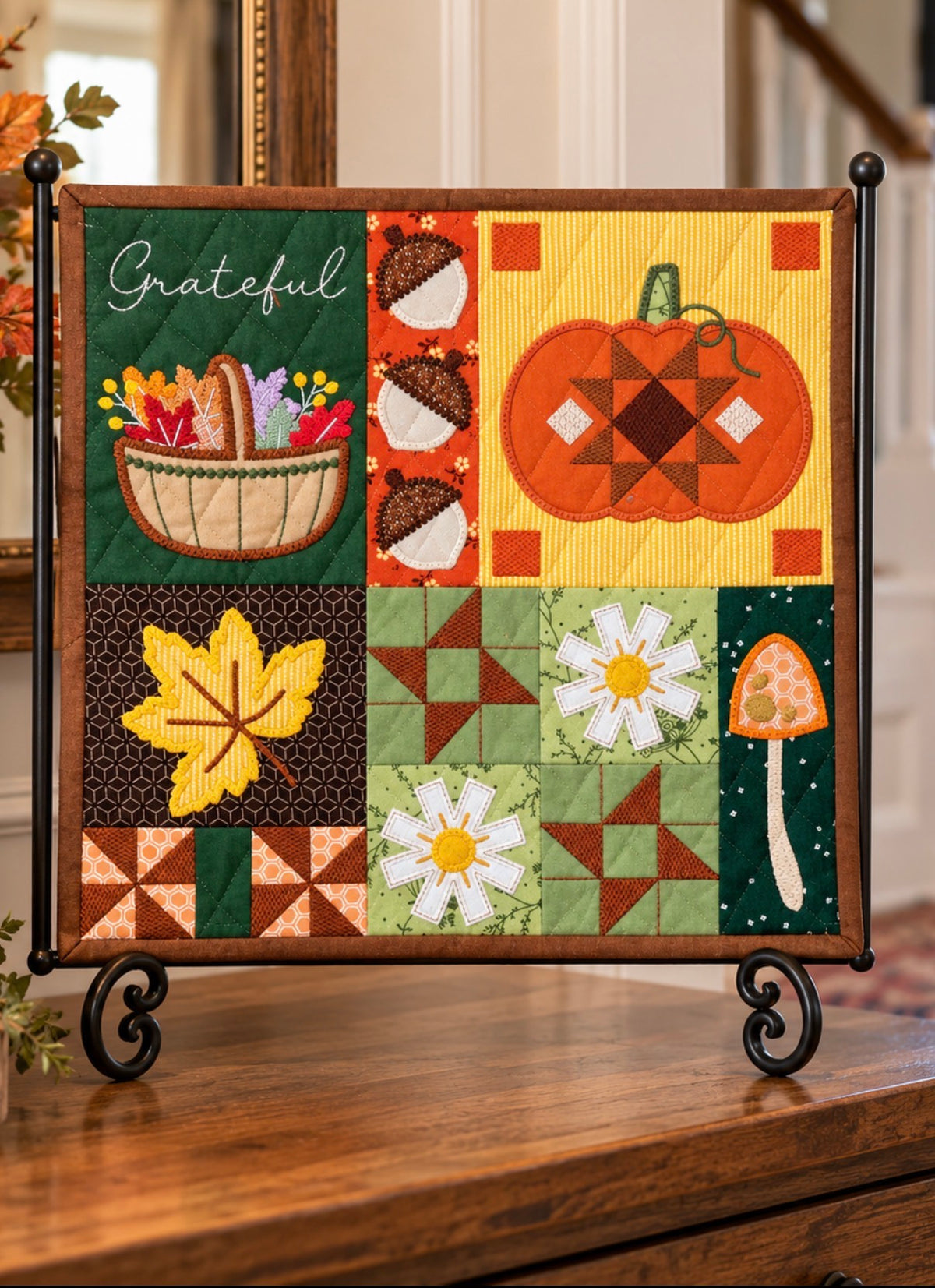 An autumn-themed fabric wall hanging quilt displayed on a black metal stand on a wooden table. The quilt features a patchwork design with seasonal motifs including a pumpkin, acorns, a leaf, daisies, a mushroom, and a basket with the word 'Grateful'.