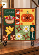 An autumn-themed fabric wall hanging quilt displayed on a black metal stand on a wooden table. The quilt features a patchwork design with seasonal motifs including a pumpkin, acorns, a leaf, daisies, a mushroom, and a basket with the word 'Grateful'.