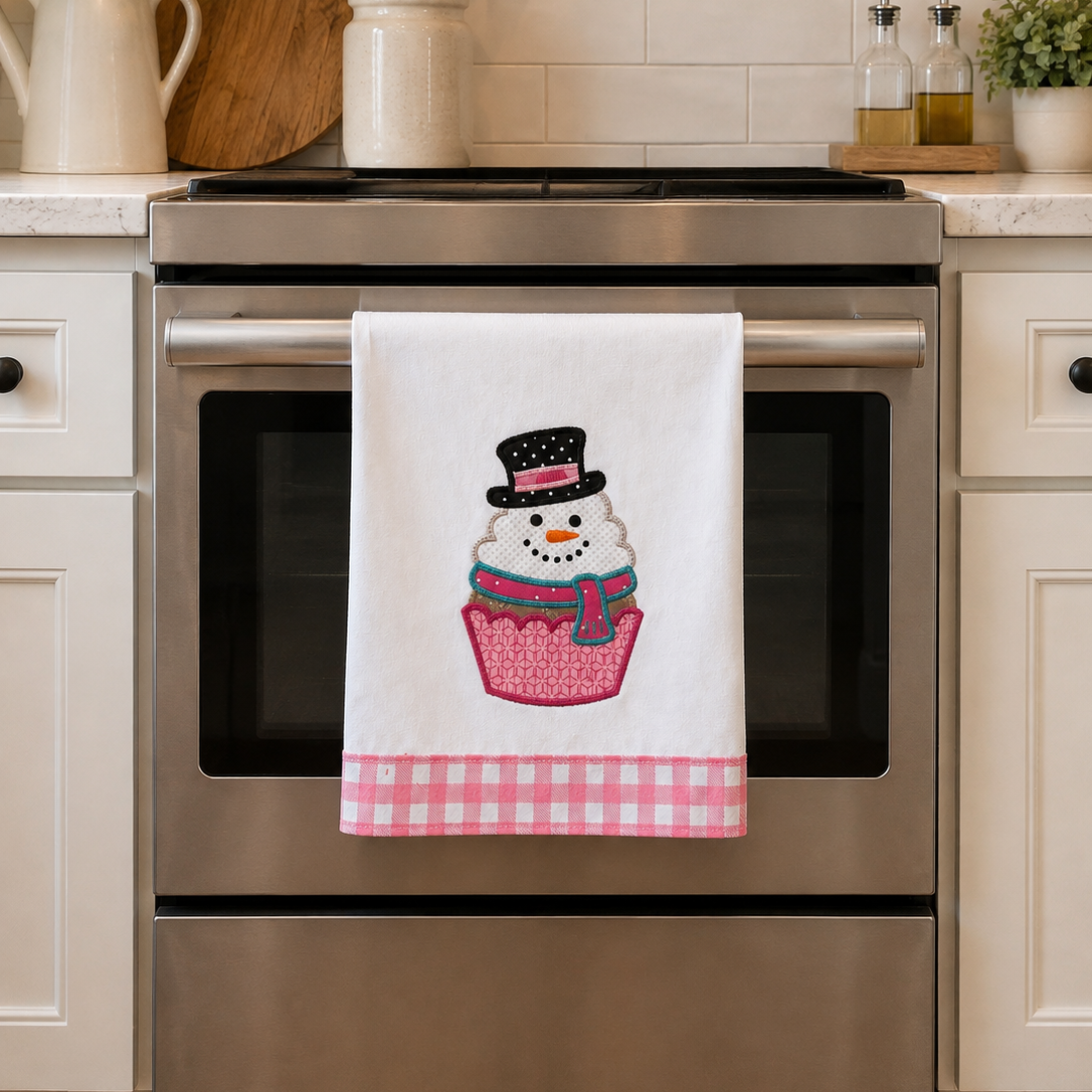A white kitchen towel hanging on a stainless steel oven handle, featuring a cupcake with a snowman wearing a black top hat and a pink gingham bottom border. The scene is set in a bright, modern kitchen with white cabinets and a tiled backsplash.
