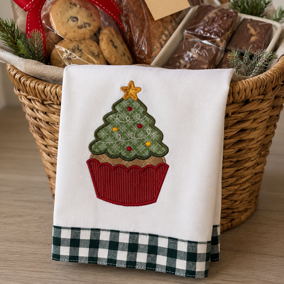 A holiday gift basket filled with cookies, chocolate bars, and wrapped treats, with a white tea towel draped over the front. The towel features an embroidered cupcake Christmas tree with a red gingham bottom border.