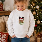 A young girl with her hair in a bun is wearing a white crewneck sweatshirt featuring a festive graphic of a reindeer wearing a Santa hat inside a green cupcake with red polka dots. The background shows a cozy Christmas setting with a decorated tree and holiday decorations.