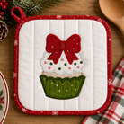 A quilted white pot holder with a red border and an applique of a cupcake topped with a red bow, shown on a wooden table with holiday decor.