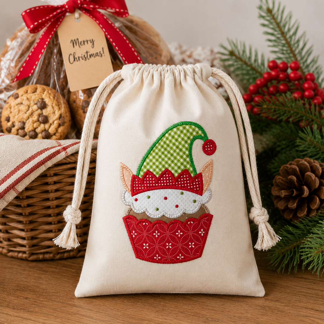 A cream fabric drawstring gift bag with an embroidered cupcake elf design, styled on a wooden table with holiday greenery, a wicker basket, and a wrapped cookie gift bag in the background.