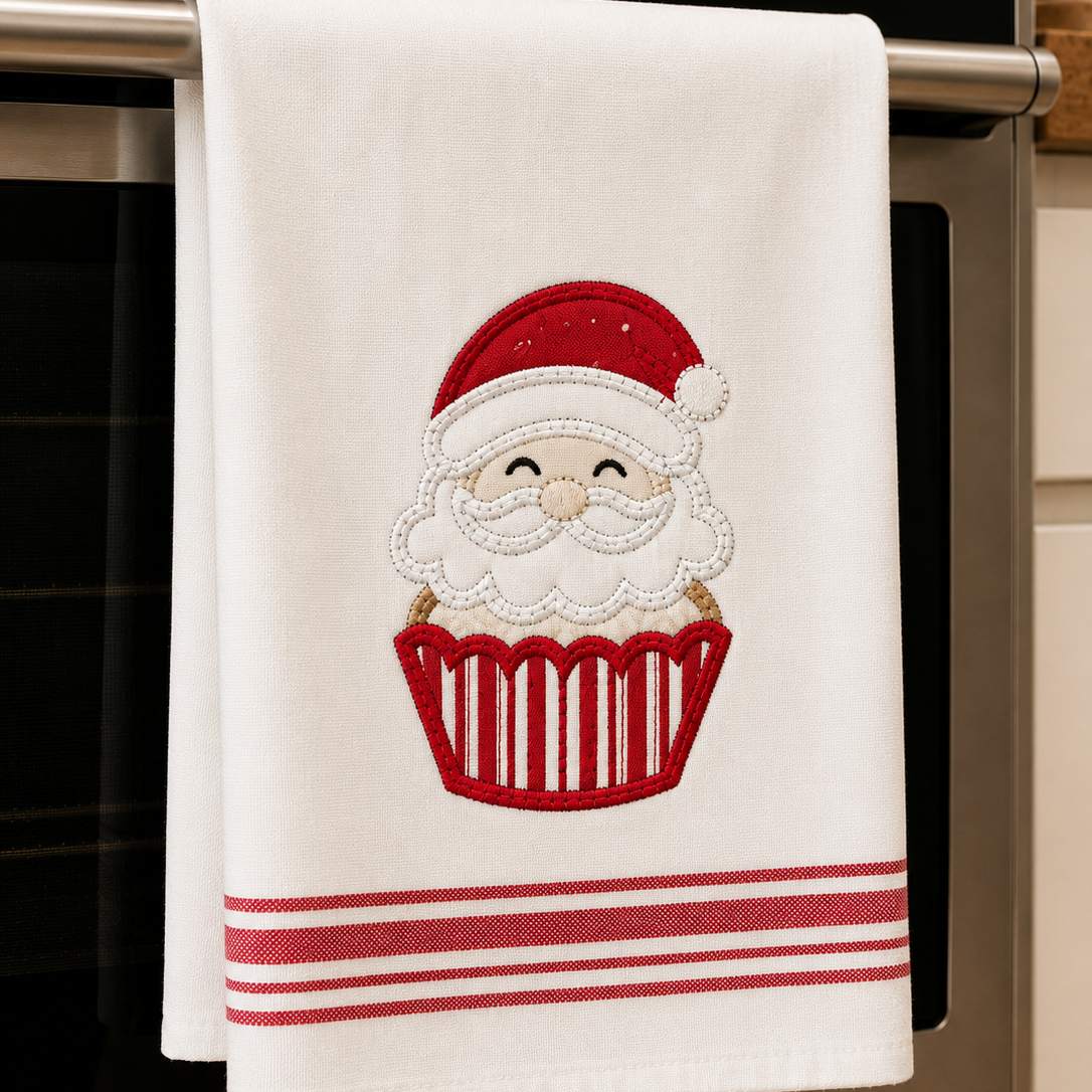A white kitchen towel hanging from a stainless steel oven handle, featuring an embroidered Santa Claus face inside a red cupcake with red stripes at the bottom.