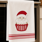 A white kitchen towel hanging from a stainless steel oven handle, featuring an embroidered Santa Claus face inside a red cupcake with red stripes at the bottom.