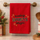 A red kitchen towel hanging on a brass towel bar, featuring the embroidered text 'making SPIRITS bright' with gold star accents, set against a white subway tile backsplash.