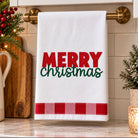 A white kitchen towel hanging on a brass towel rack, featuring the text 'MERRY Christmas' in red and green, with a red and white gingham stripe at the bottom. The towel is displayed in a decorated kitchen setting with holiday greenery, a cutting board, and a marble countertop.