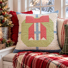 A square decorative throw pillow with a quilted holiday gift design in red, green, and blue, styled on a cozy window seat with a red plaid blanket and a Christmas tree nearby.