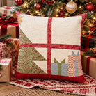 A square quilted throw pillow with a Christmas-themed patchwork design featuring a tree, gifts, and a cross, styled on a rug in front of a decorated Christmas tree and wrapped presents.