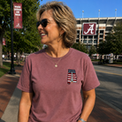 Woman wearing a maroon t-shirt with a logo, standing on a sidewalk with a stadium in the background.
