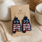 Pair of earrings on a card with 'Made with Love' text, placed on a textured surface with a plant and cup in the background.