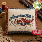 Embroidered pillow with 'America 250 Sew Blessed 1776-2026' design on a wooden surface with sewing tools.