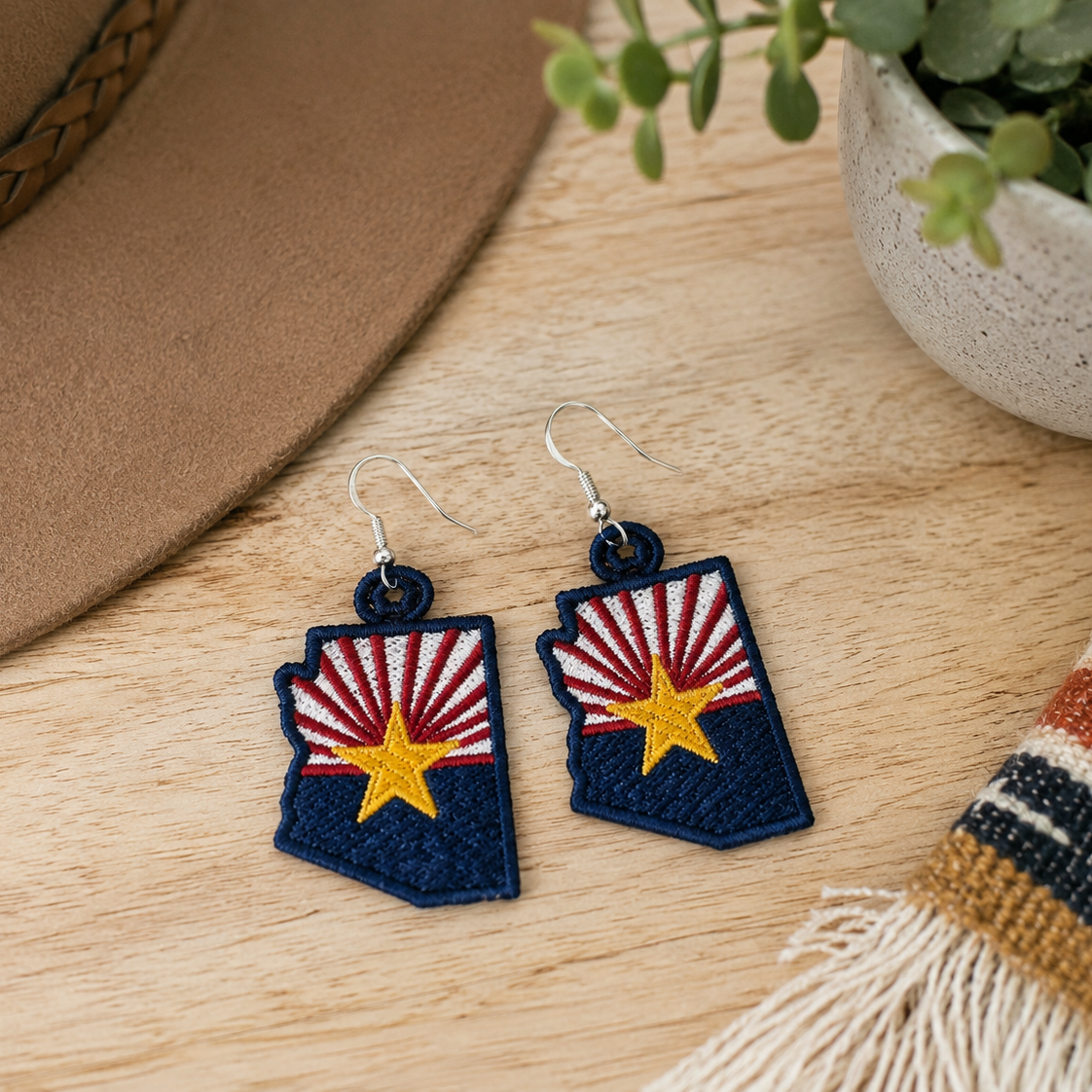 Arizona-shaped earrings with a flag design on a wooden surface with a hat and plant in the background.