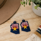 Arizona-shaped earrings with a flag design on a wooden surface with a hat and plant in the background.