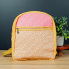 A small quilted crossbody bag with a pink top panel and a yellow floral patterned front pocket, displayed on a wooden surface next to a small potted plant against a dark gray background.